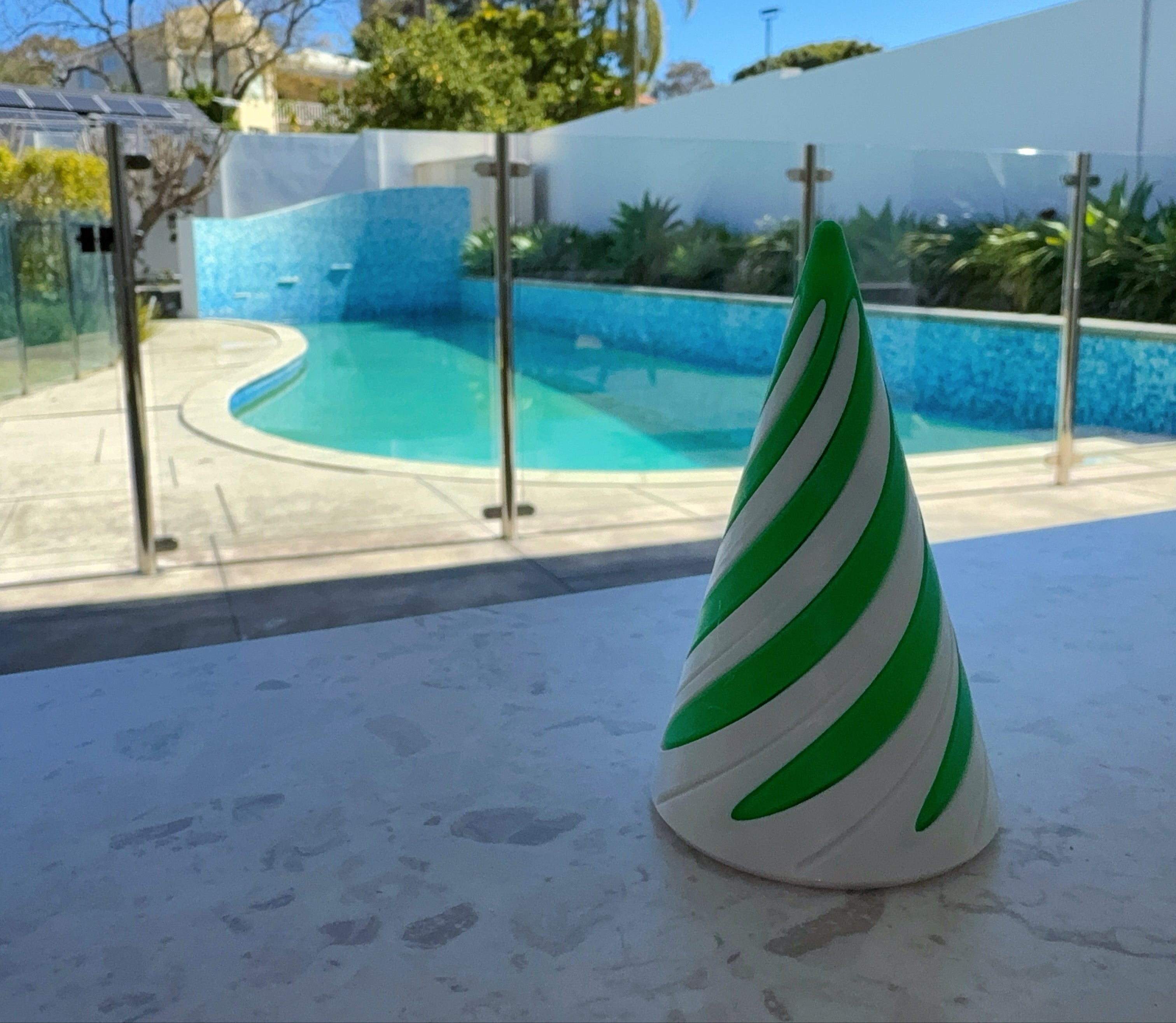 Green and white Sprially striped cone-shaped fidget toy on a table with a pool and garden in the background.